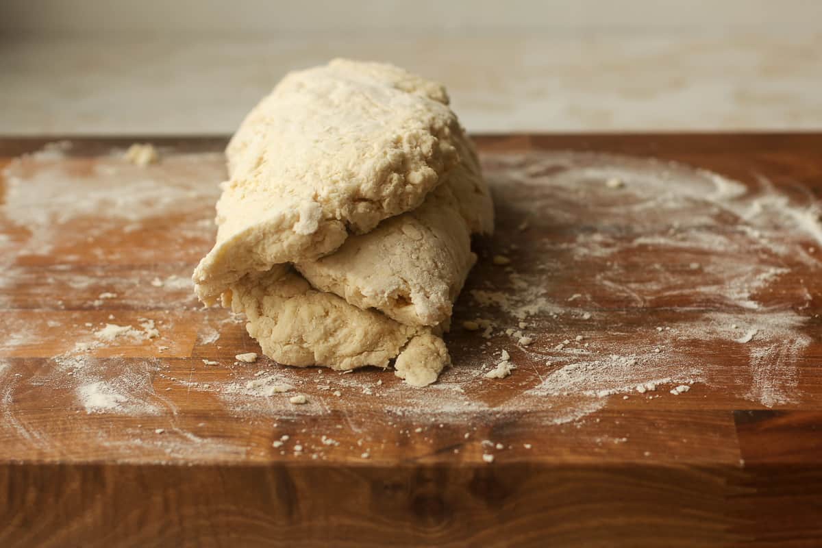 Hands folding a rectangle of biscuit dough on a lightly floured countertop, with a bench scraper nearby