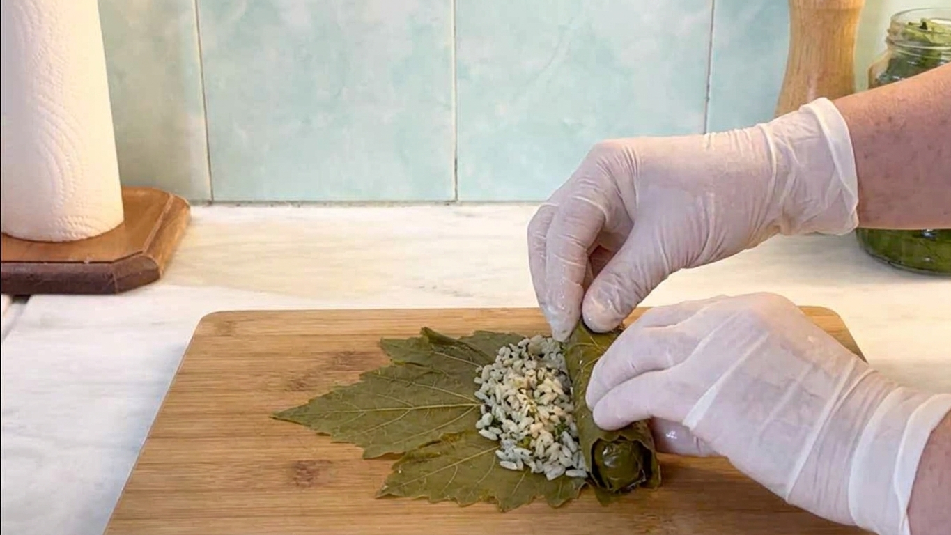 Hands folding and rolling a grape leaf around a spoonful of herbed rice filling on a wooden cutting board