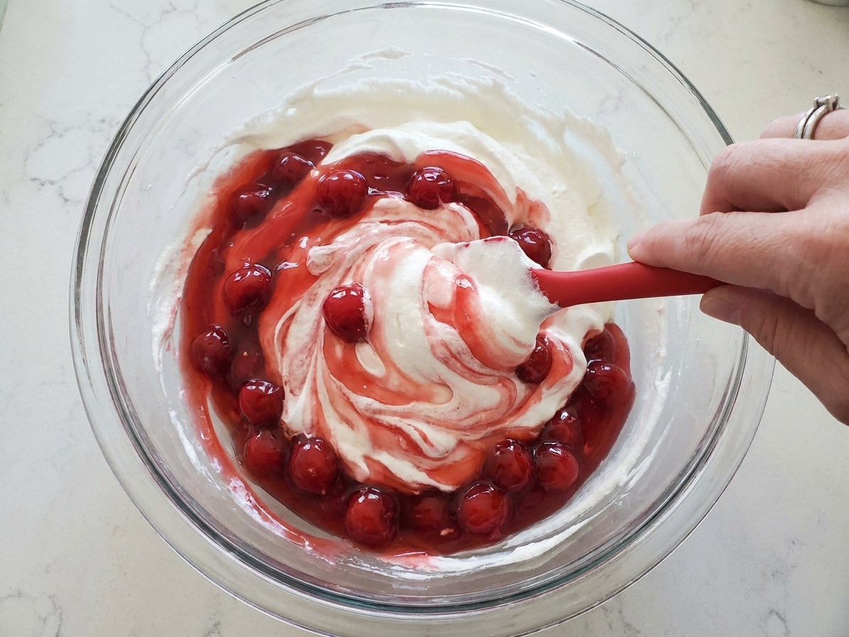 Hands folding whipped topping and cherry pie filling together in a large mixing bowl with a rubber spatula