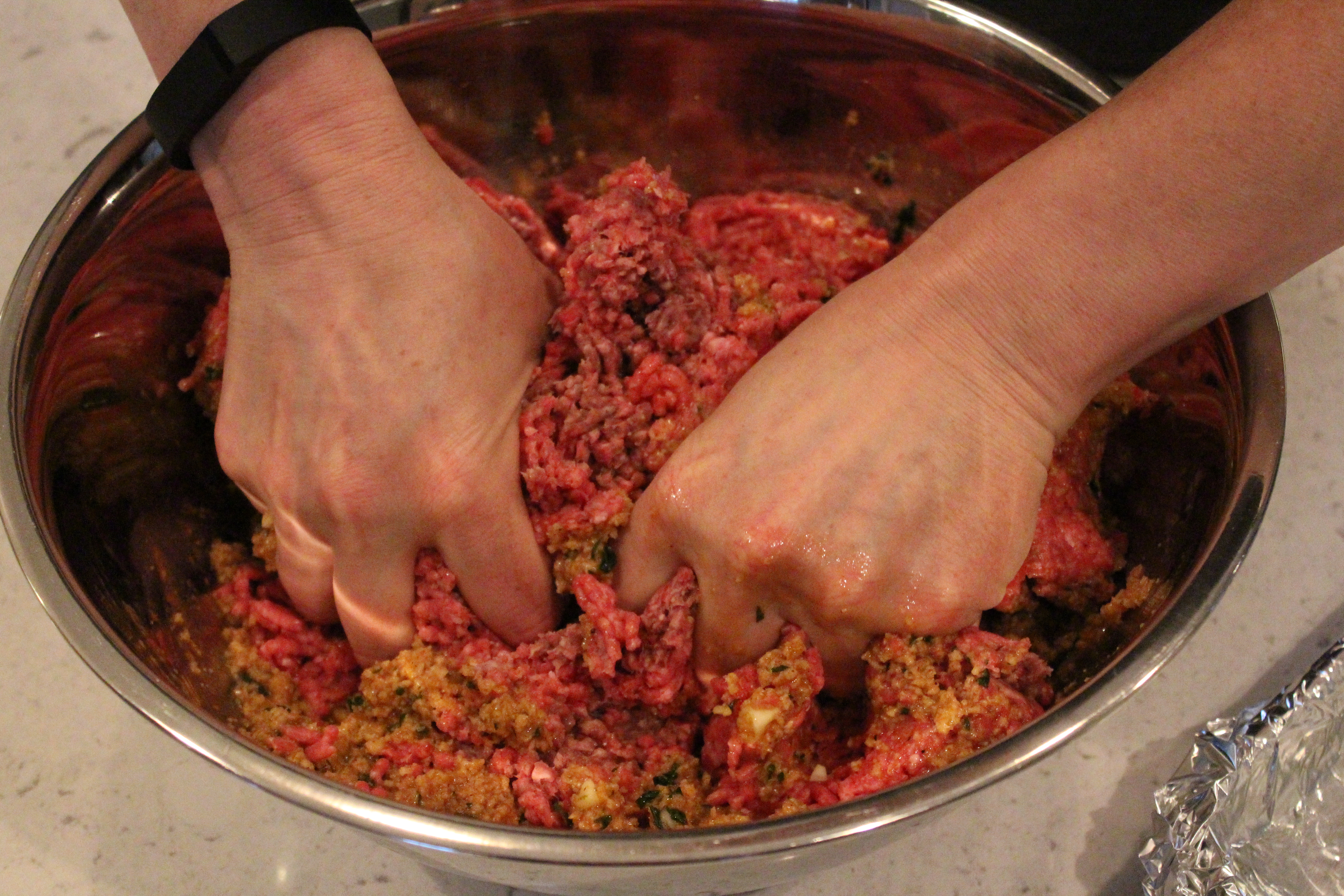 Hands gently mixing ground beef, sautéed onions, breadcrumbs, and eggs in a large glass bowl on a home kitchen counter