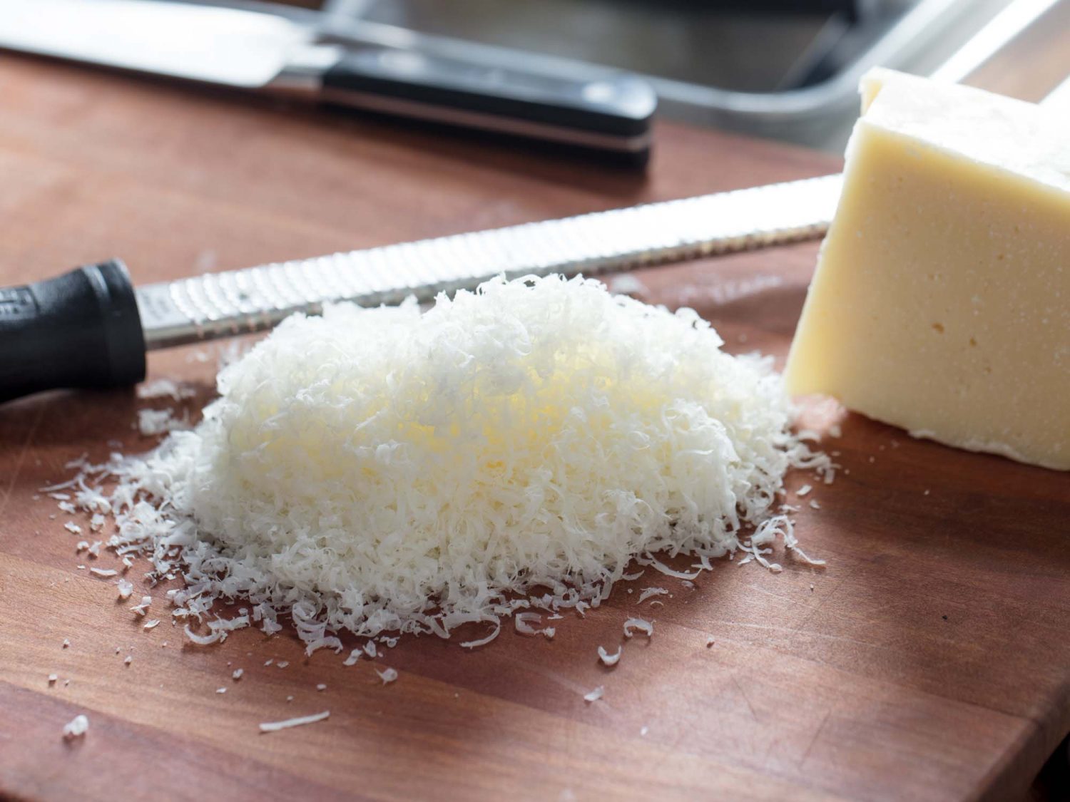 Hands grating Pecorino Romano with a microplane over a wooden cutting board beside a skillet of red tomato sauce on a home kitchen counter