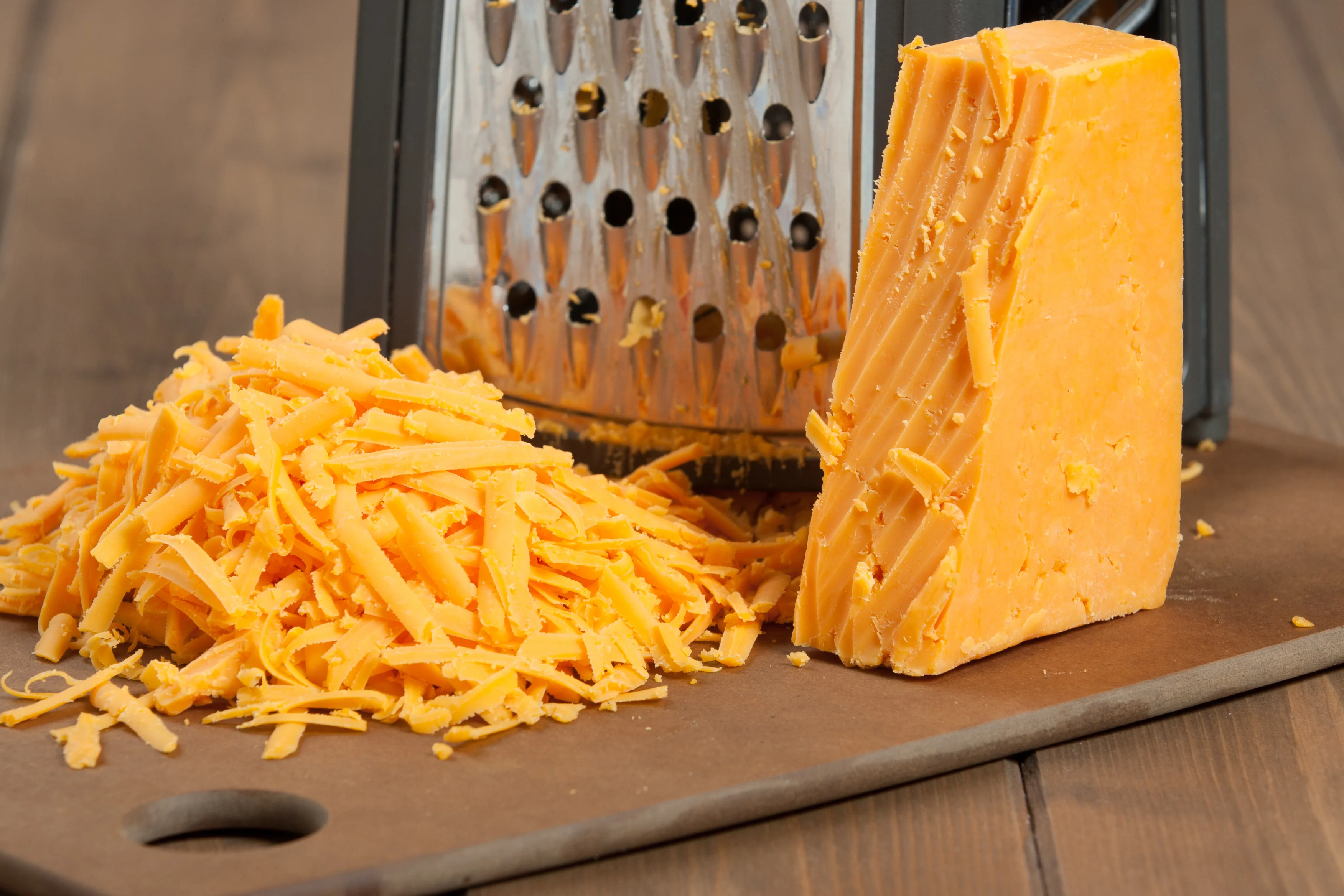 Hands grating a block of sharp cheddar cheese over a bowl on a kitchen counter with a box grater