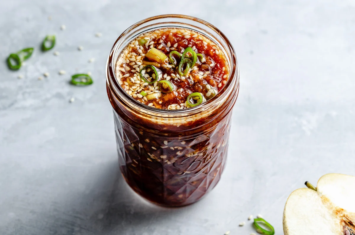 Hands grating a ripe pear over a bowl on a kitchen counter, with soy sauce and sesame oil nearby