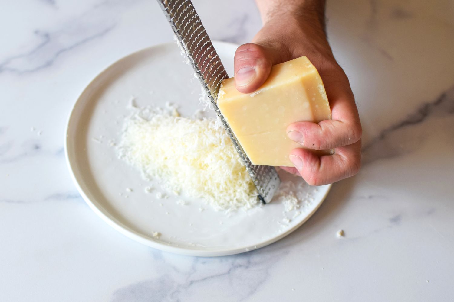 Hands grating a wedge of Parmesan with a microplane over a small bowl on a bright kitchen counter