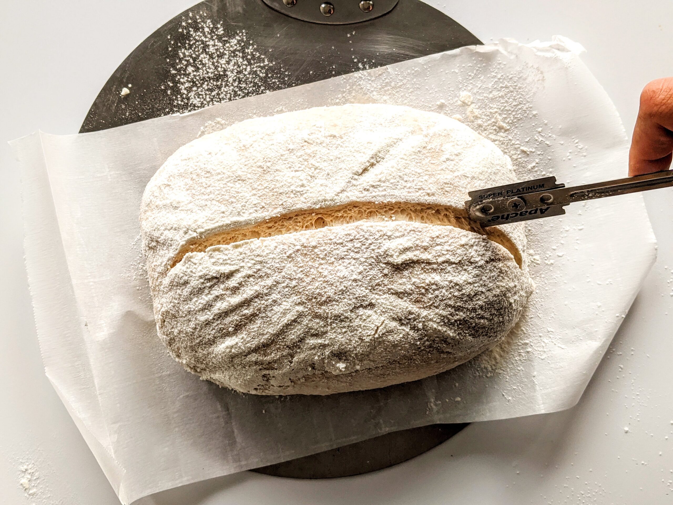 Hands holding a bread lame scoring a proofed sourdough loaf on parchment paper on a wooden counter