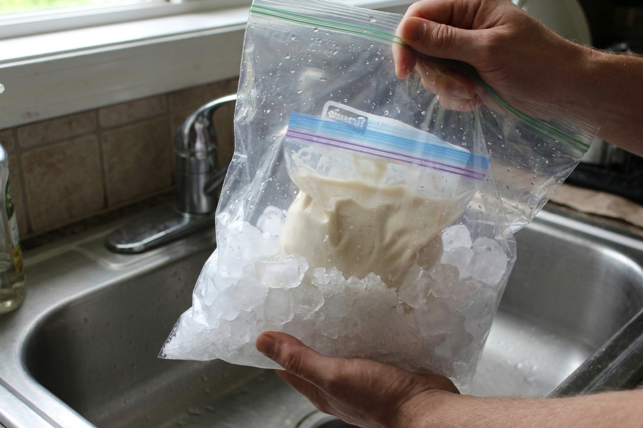 Hands holding a large zip-top bag filled with ice and salt while shaking a smaller bag of ice cream base inside over a kitchen sink