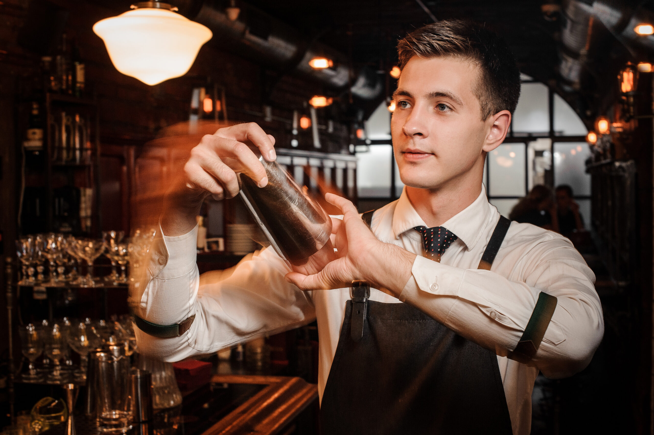 Hands holding a metal cocktail shaker on a kitchen counter mid dry shake, with lemon halves and a jigger nearby