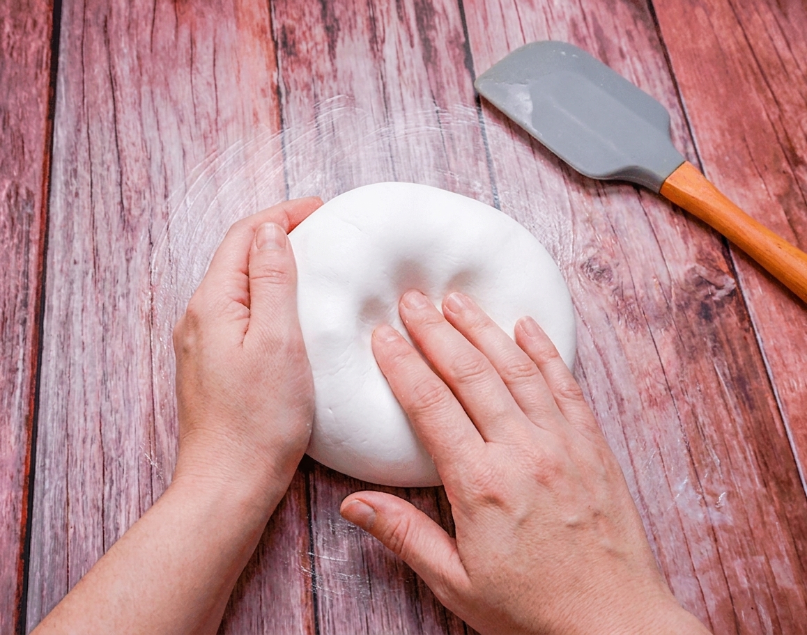 Hands kneading a smooth ball of white fondant on a lightly greased kitchen counter with a silicone spatula nearby