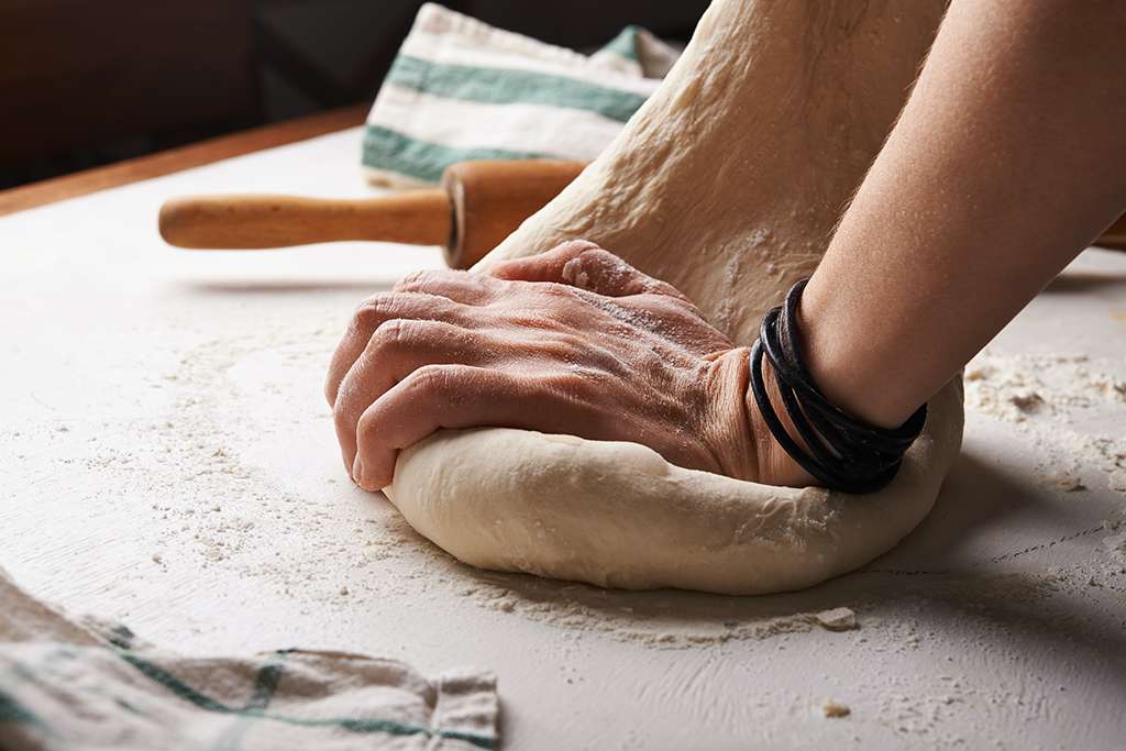 Hands kneading pizza dough on a lightly floured countertop with a bench scraper resting nearby