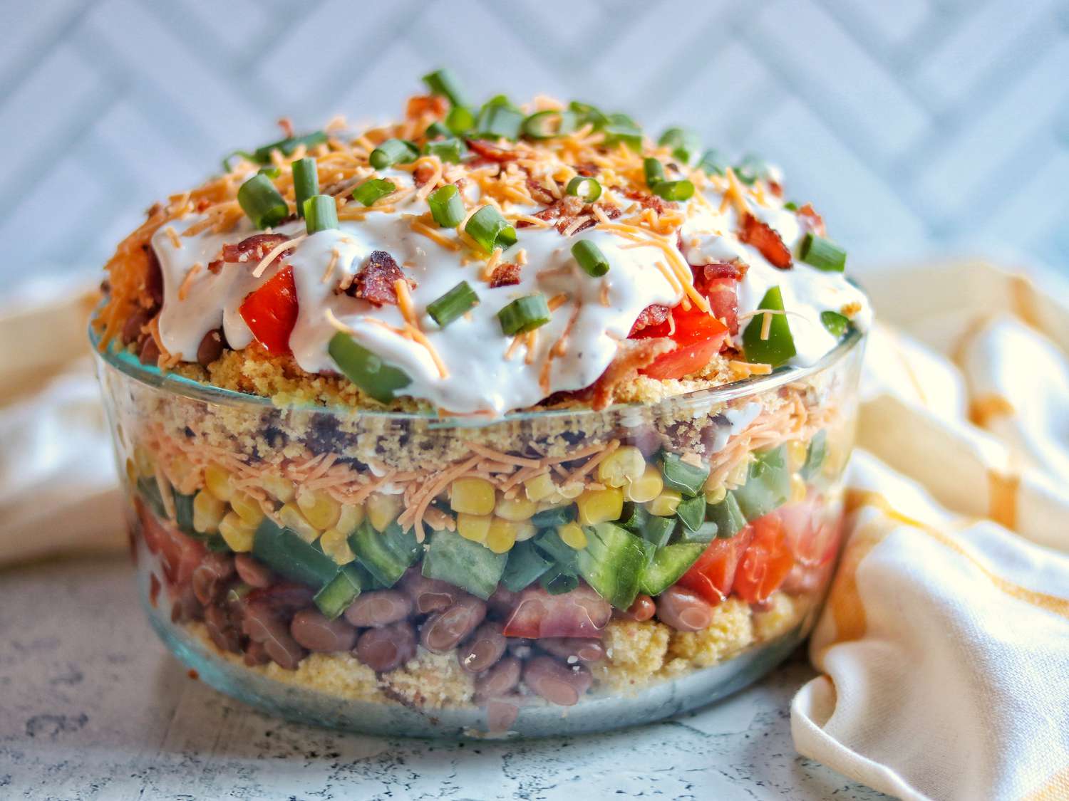Hands layering toasted cornbread cubes, tomatoes, beans, and shredded cheese into a clear glass trifle dish on a kitchen counter