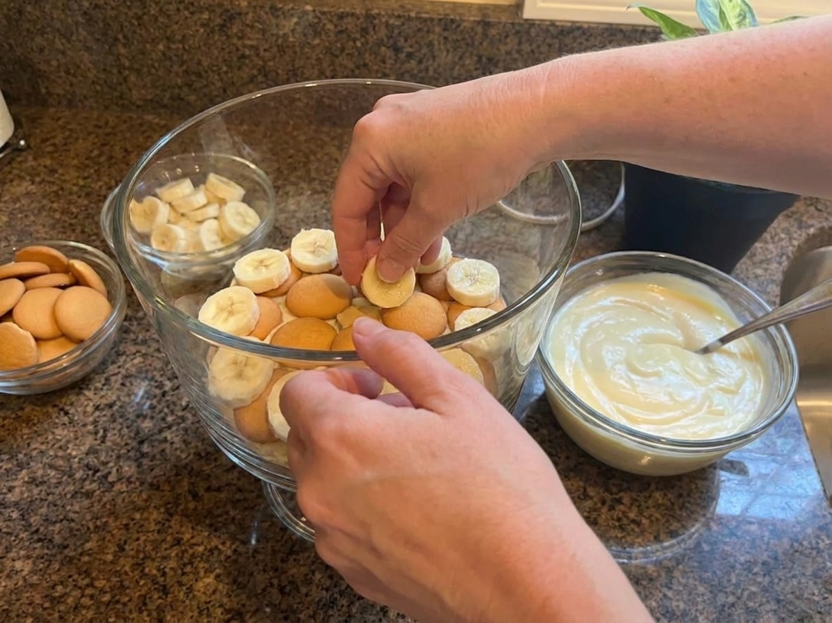 Hands layering vanilla wafers and banana slices into a glass trifle dish on a kitchen counter before adding pudding