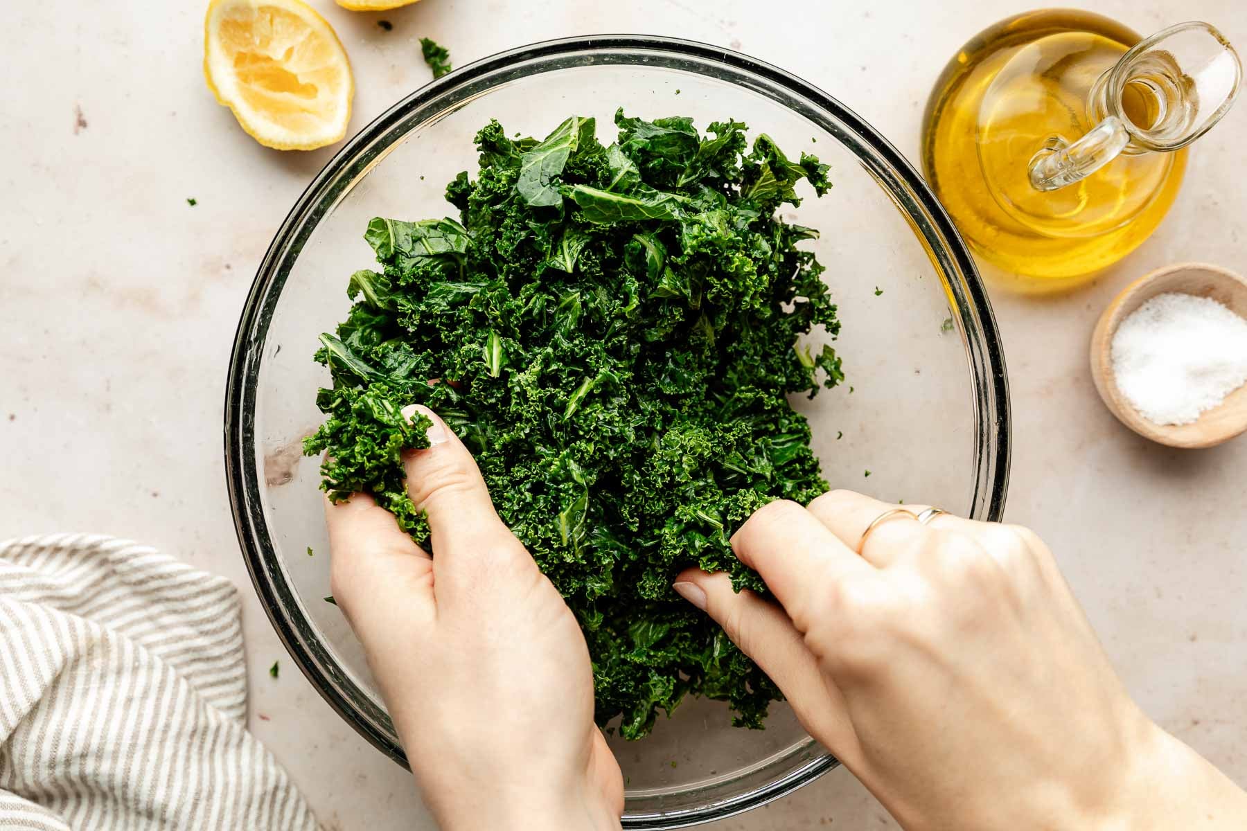 Hands massaging thinly sliced kale with a pinch of salt in a large mixing bowl on a light stone kitchen counter
