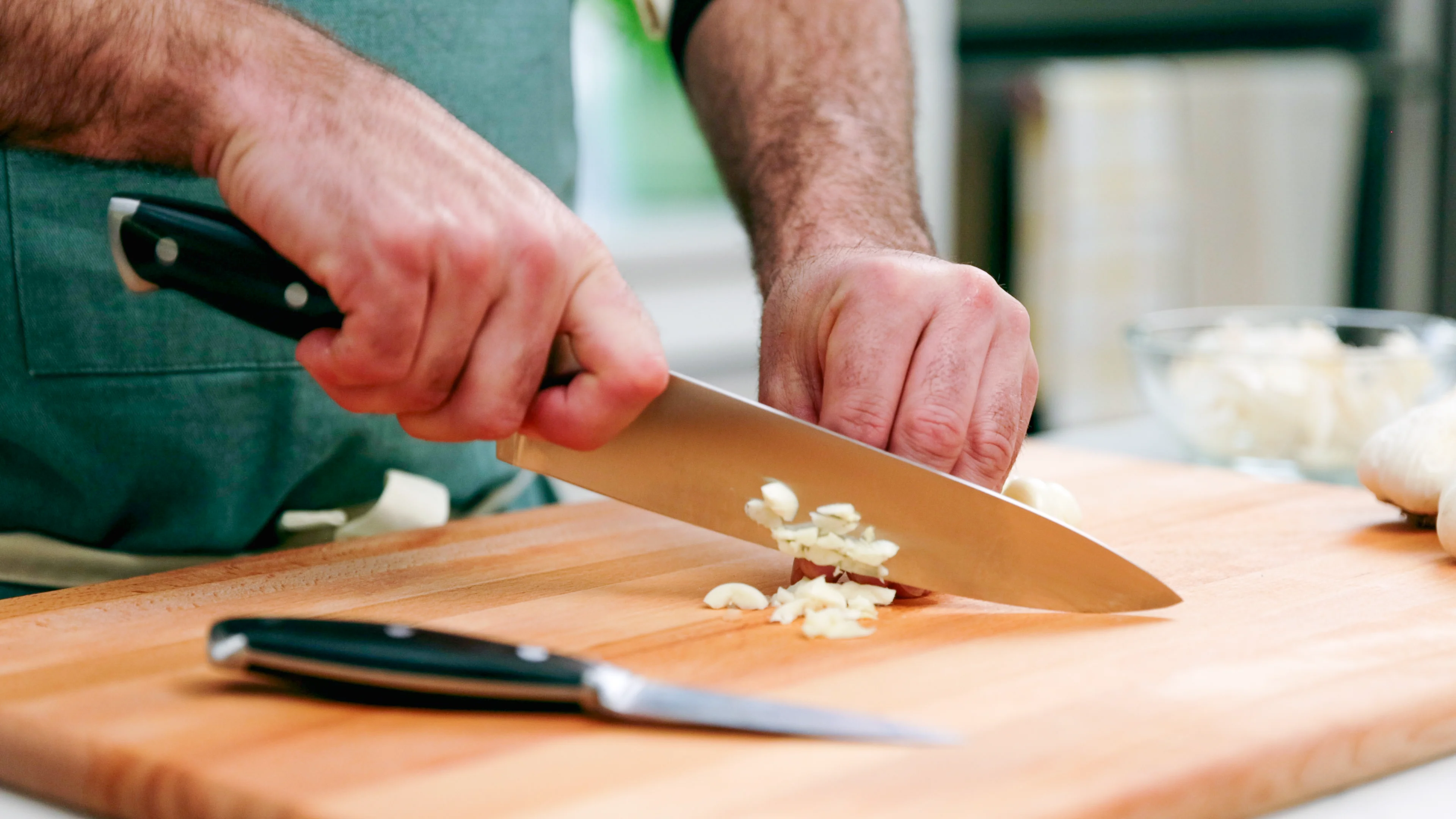 Hands mincing fresh garlic with a chef's knife on a wooden cutting board on a home kitchen counter