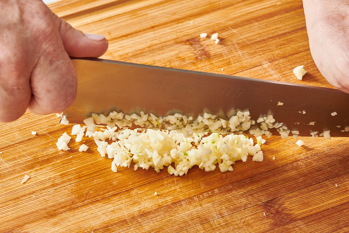 Hands mincing garlic on a wooden cutting board beside trimmed green beans and a small bowl of grated ginger in a bright home kitchen