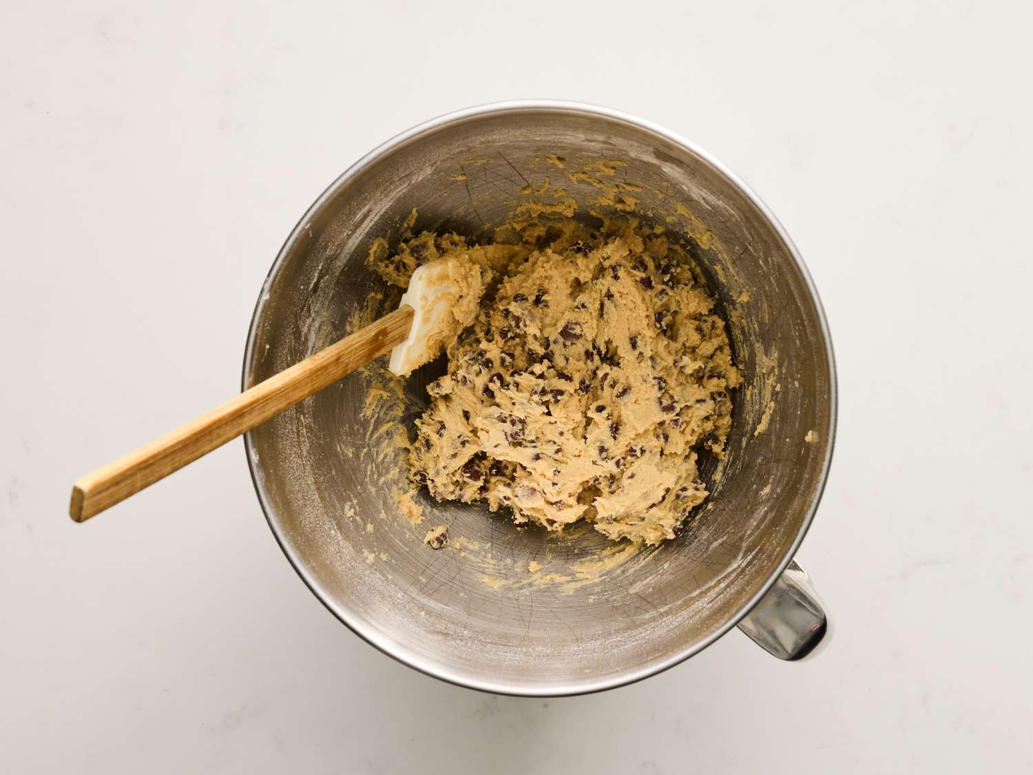 Hands mixing edible cookie dough in a glass bowl with a silicone spatula on a kitchen counter under warm light