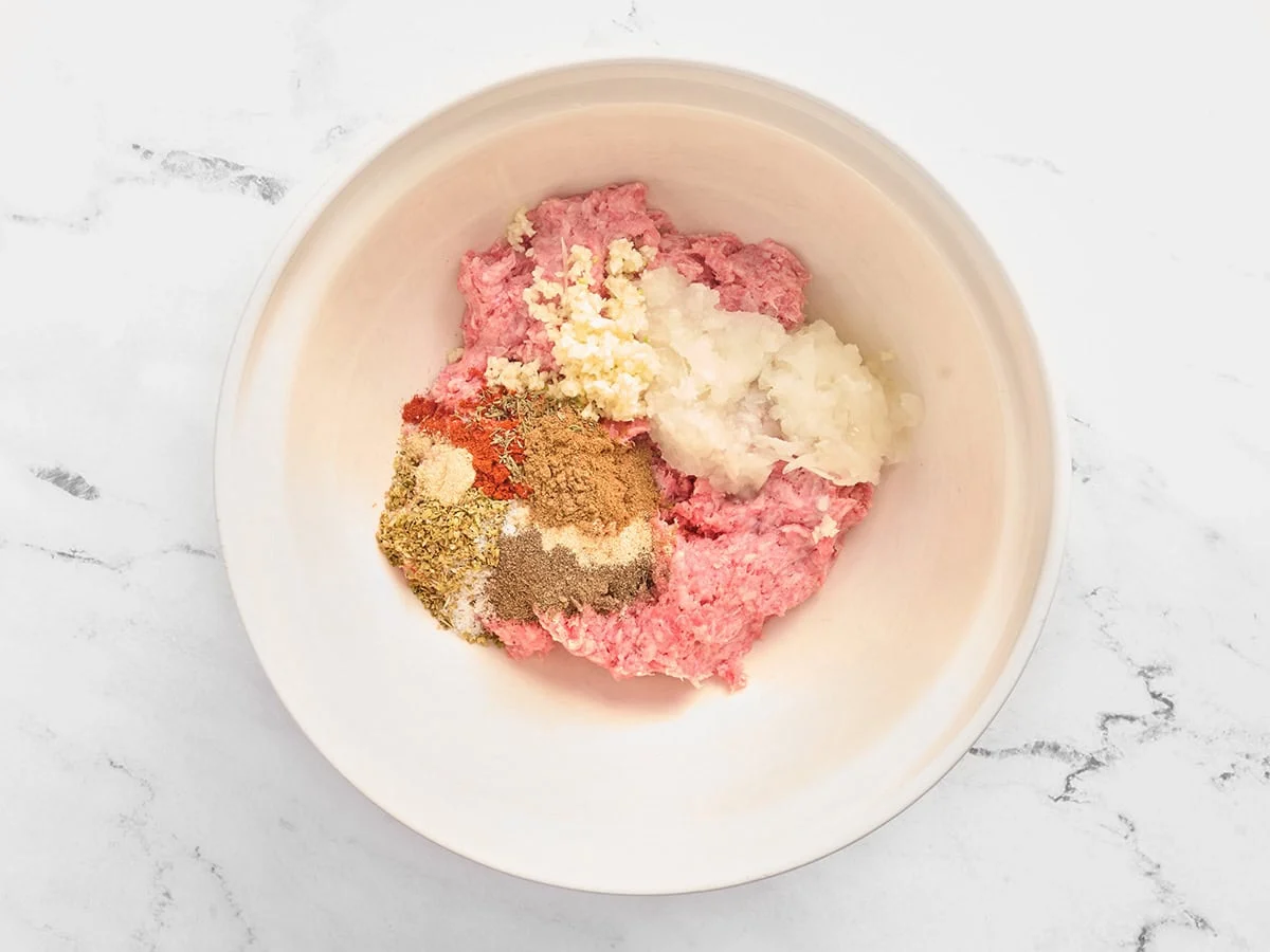Hands mixing ground beef and lamb with grated onion and spices in a large stainless steel mixing bowl on a kitchen counter