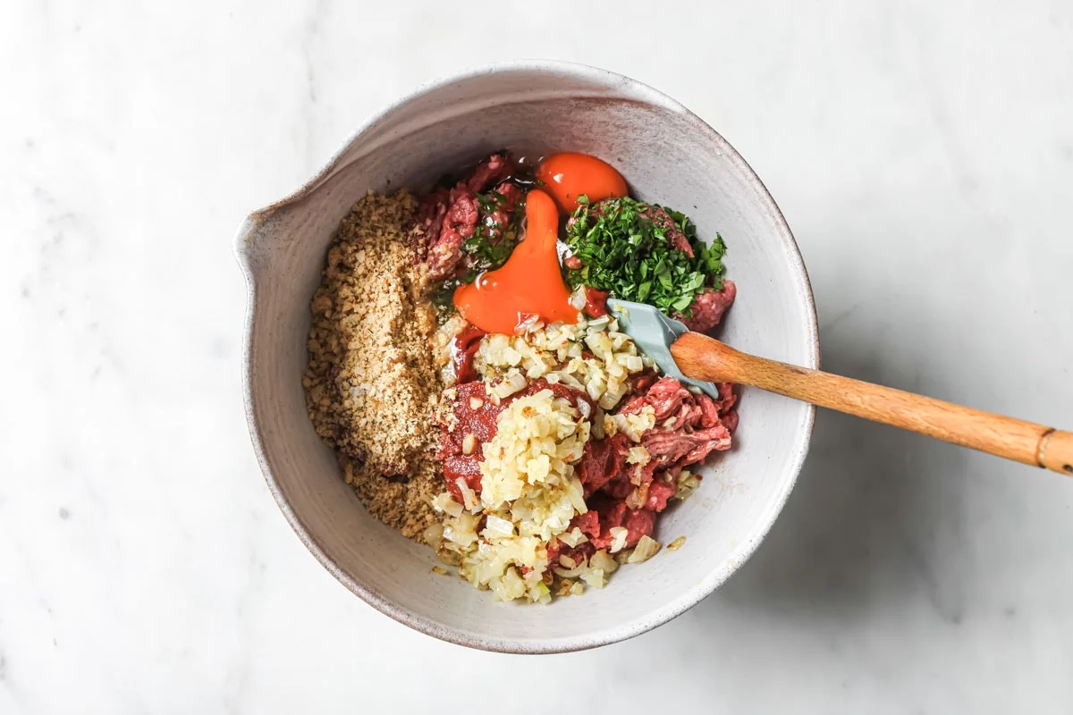 Hands mixing ground beef with eggs, almond flour, parmesan, onion, and seasonings in a large glass bowl on a kitchen counter