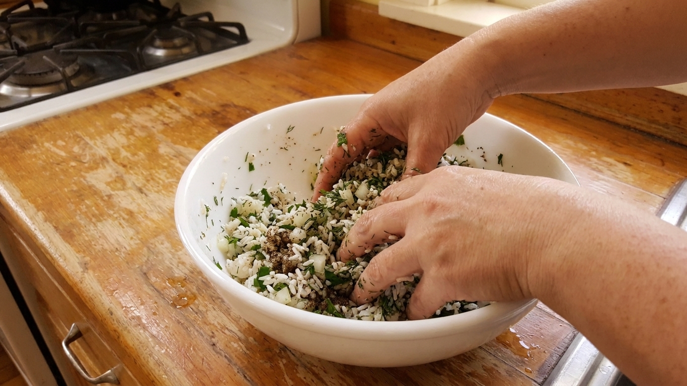 Hands mixing rice, chopped herbs, onion, and spices in a large white bowl on a kitchen counter