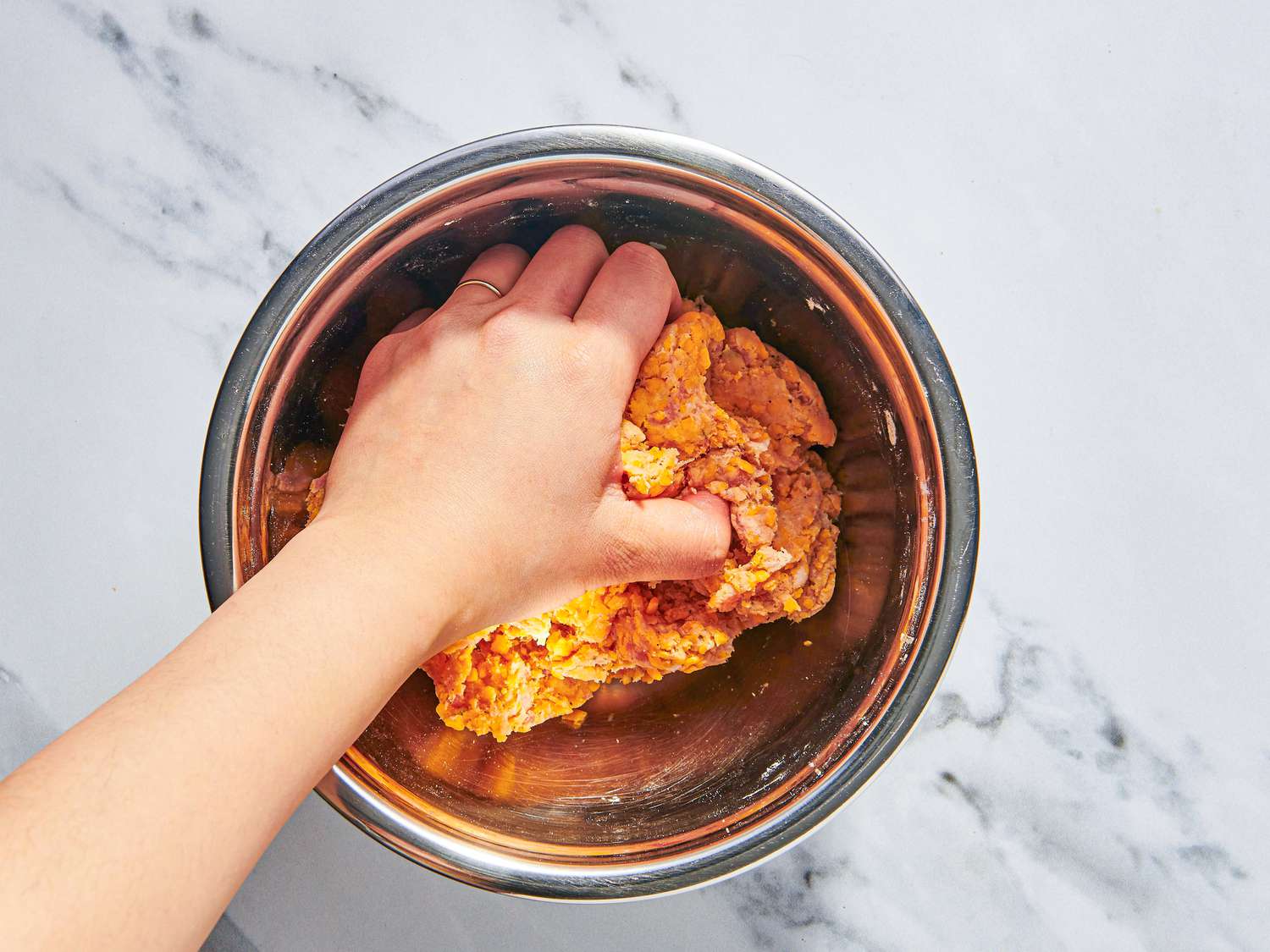 Hands mixing sausage, Bisquick, shredded cheddar, and milk in a large glass mixing bowl on a kitchen counter