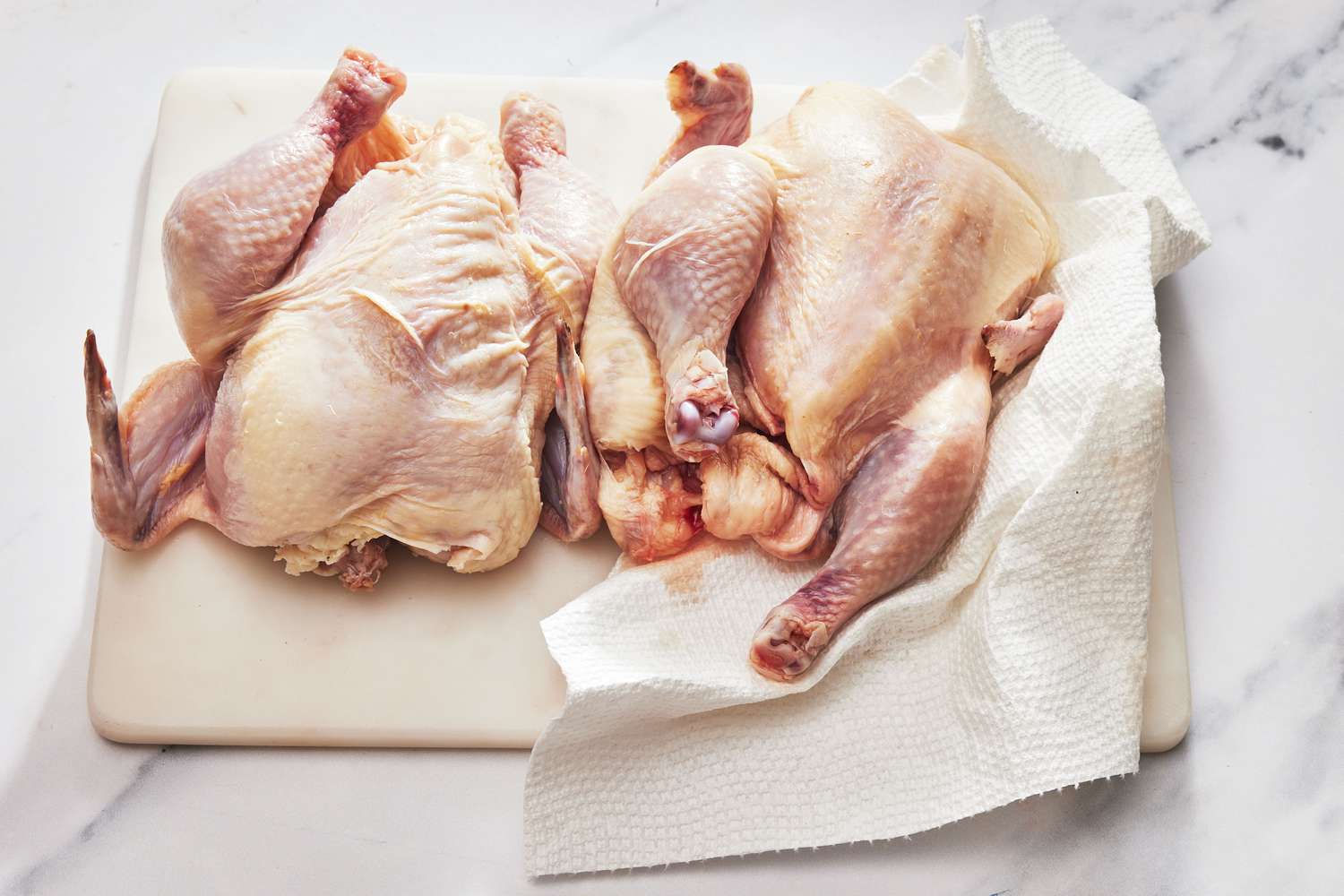 Hands patting a raw Cornish game hen dry with paper towels on a cutting board beside a roasting pan
