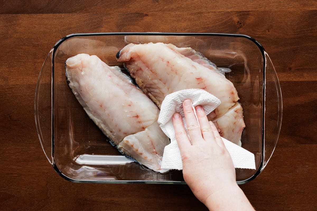 Hands patting raw fish fillets dry with paper towels before seasoning