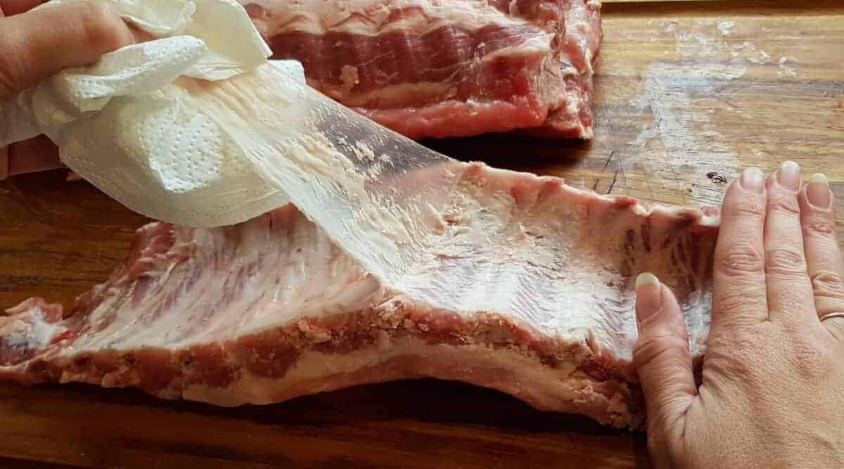 Hands peeling the thin membrane from the back of a rack of pork ribs using a paper towel on a cutting board