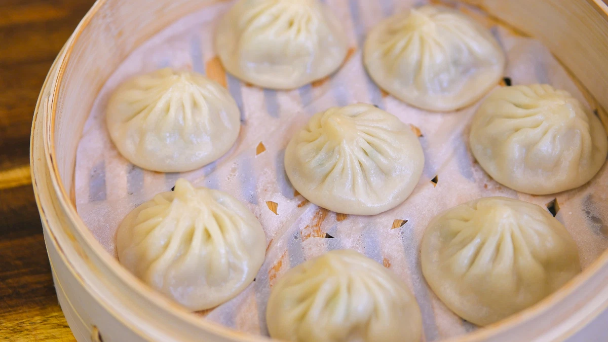 Hands pleating a xiao long bao dumpling on a floured wooden board, with other wrappers and filling nearby.