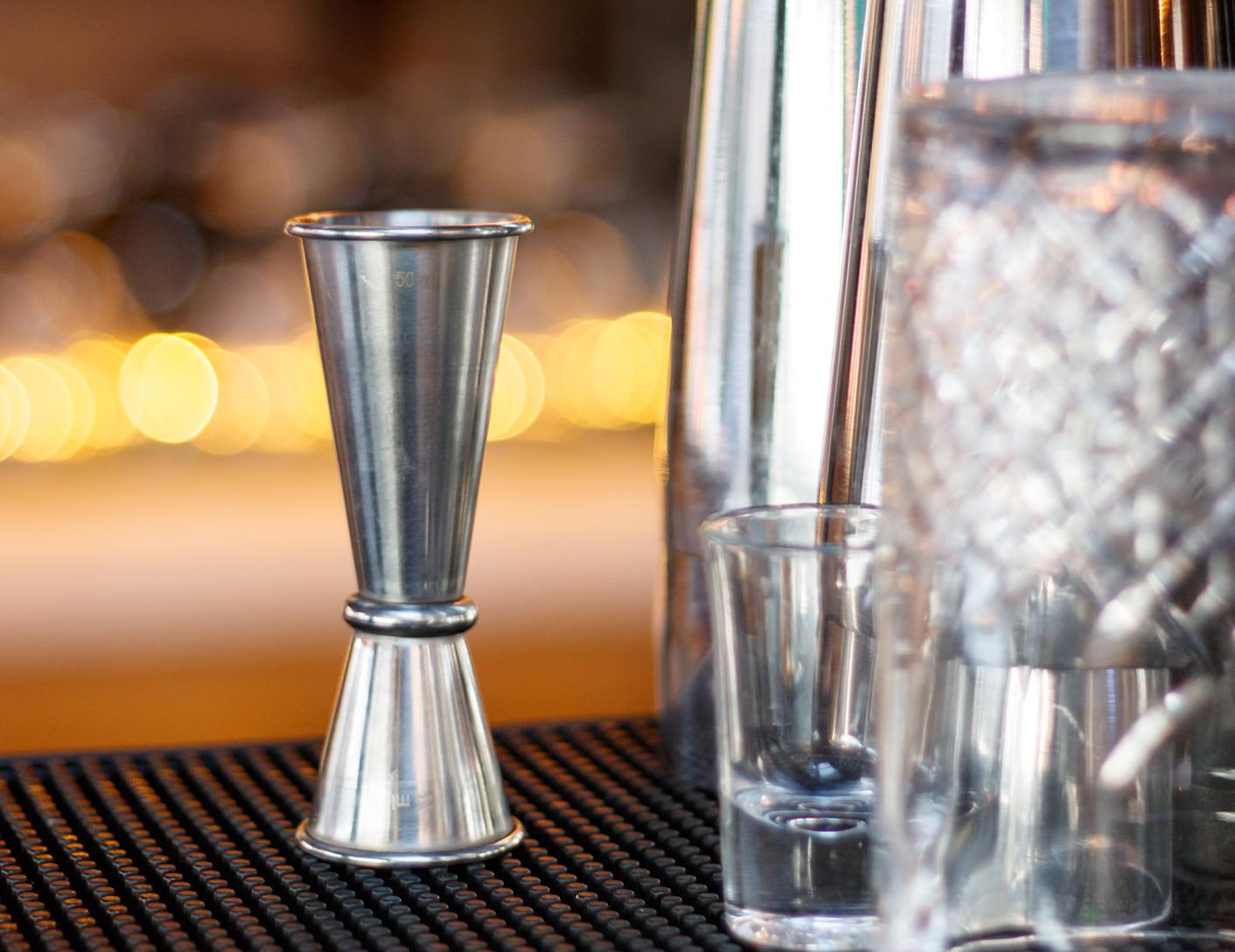 Hands pouring clear vodka from a jigger into a tall glass filled with ice on a kitchen counter