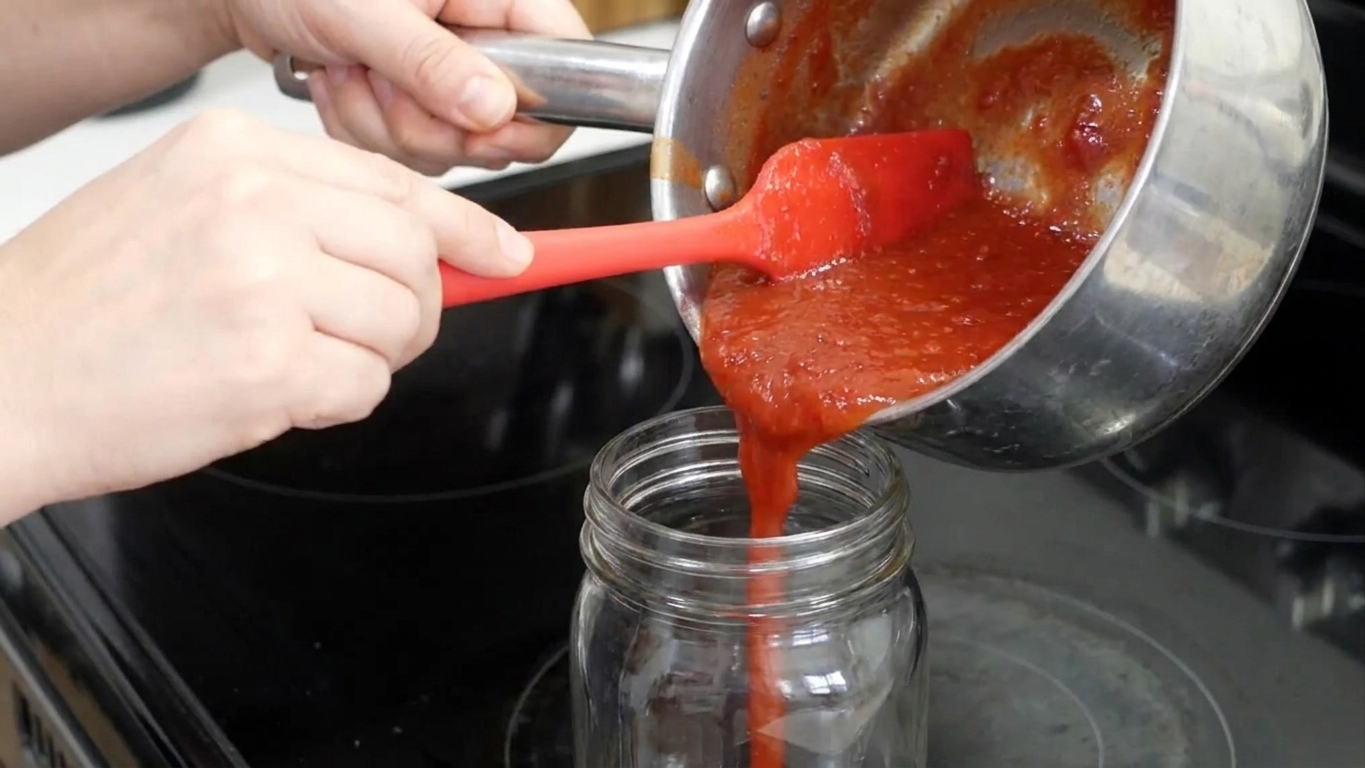 Hands pouring cooled homemade ketchup from a saucepan into a clean glass jar on a kitchen counter