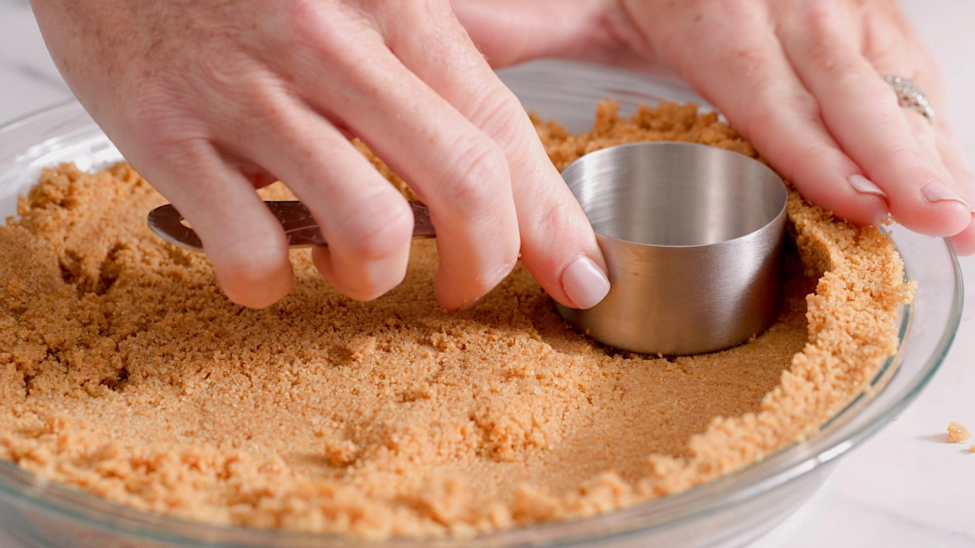 Hands pressing a buttery graham cracker crumb crust into a parchment-lined 9x13-inch baking pan on a kitchen counter