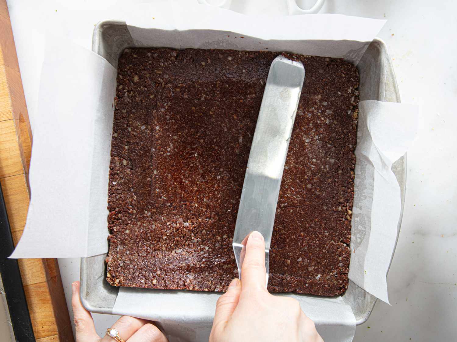 Hands pressing a chocolate coconut crumb mixture firmly into a parchment-lined square baking pan on a kitchen counter