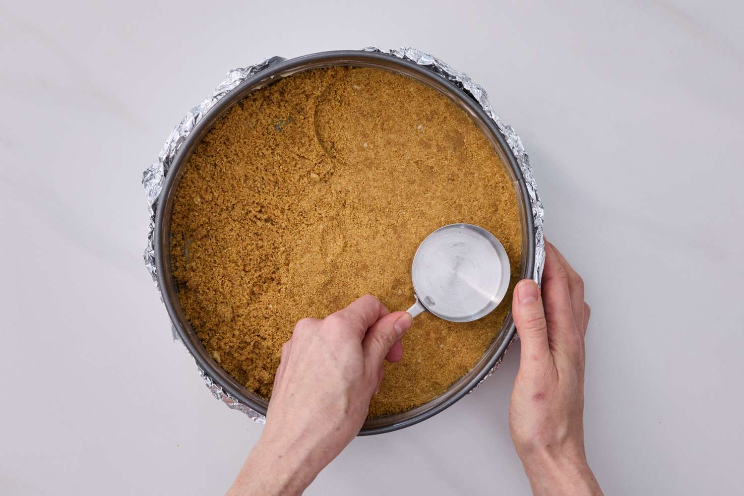 Hands pressing buttery graham cracker crumbs into the bottom and up the sides of a springform pan on a kitchen counter