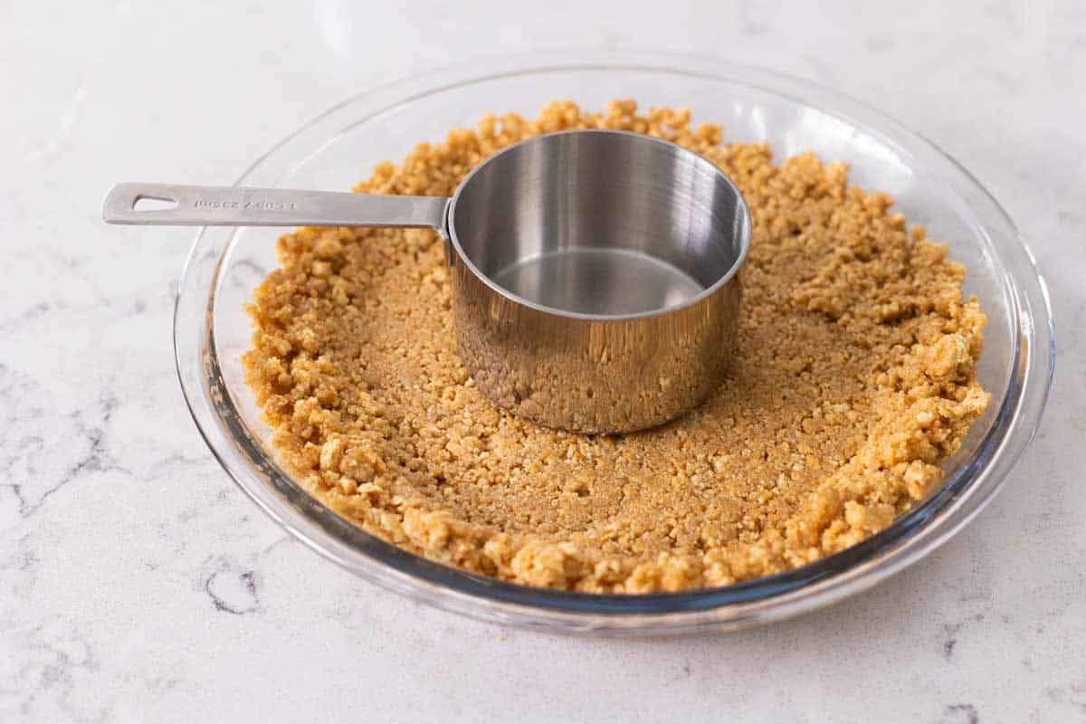 Hands pressing graham cracker crust mixture firmly into a parchment-lined square baking pan on a kitchen counter