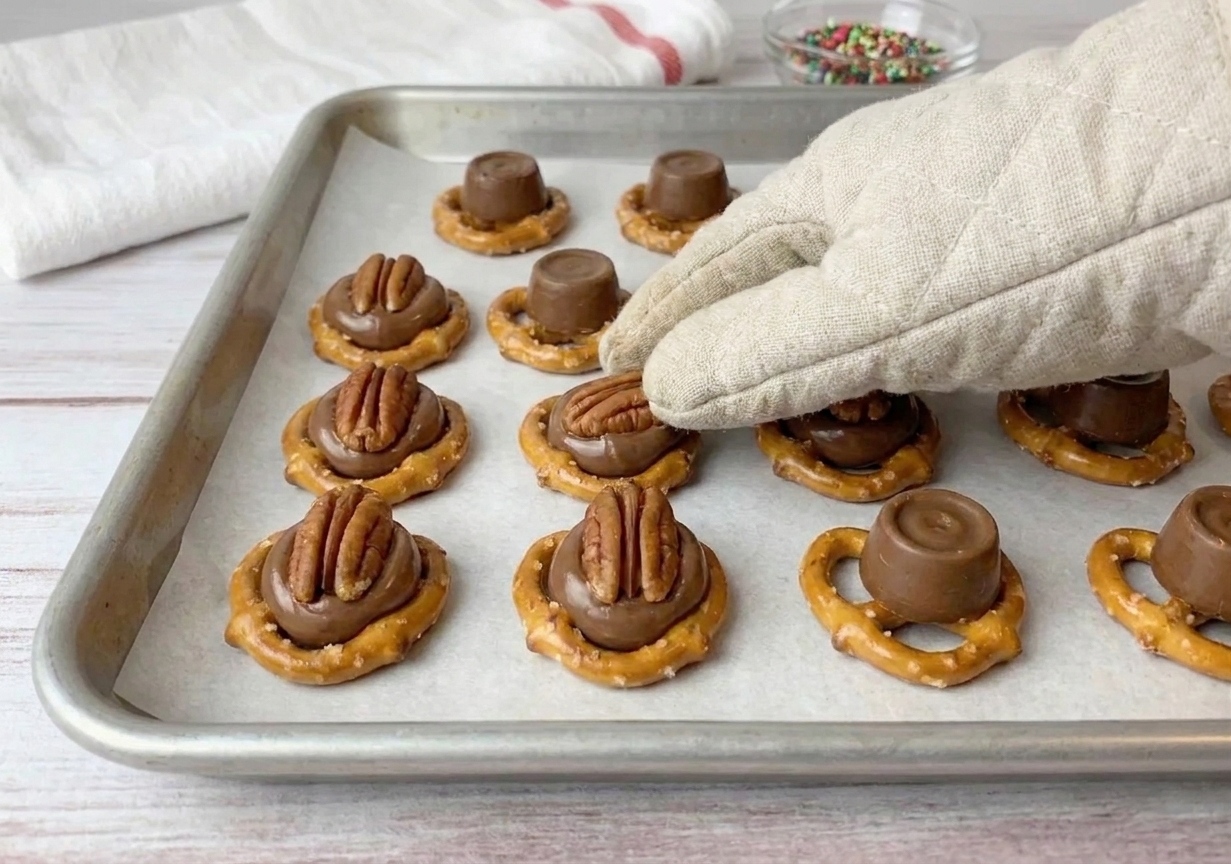 Hands pressing pecan halves into softened Rolo-topped pretzels on a parchment-lined baking sheet, creating neat bite-sized candy clusters