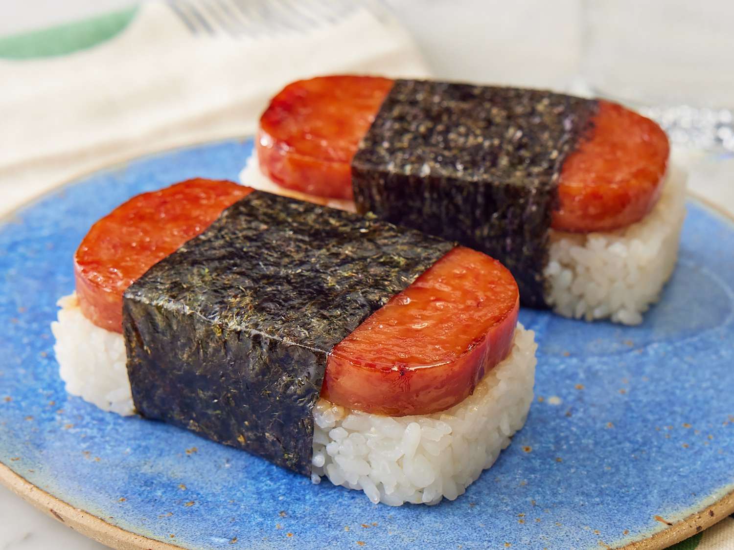 Hands pressing seasoned sushi rice into a musubi mold on a cutting board, with nori strips and seared Spam slices nearby