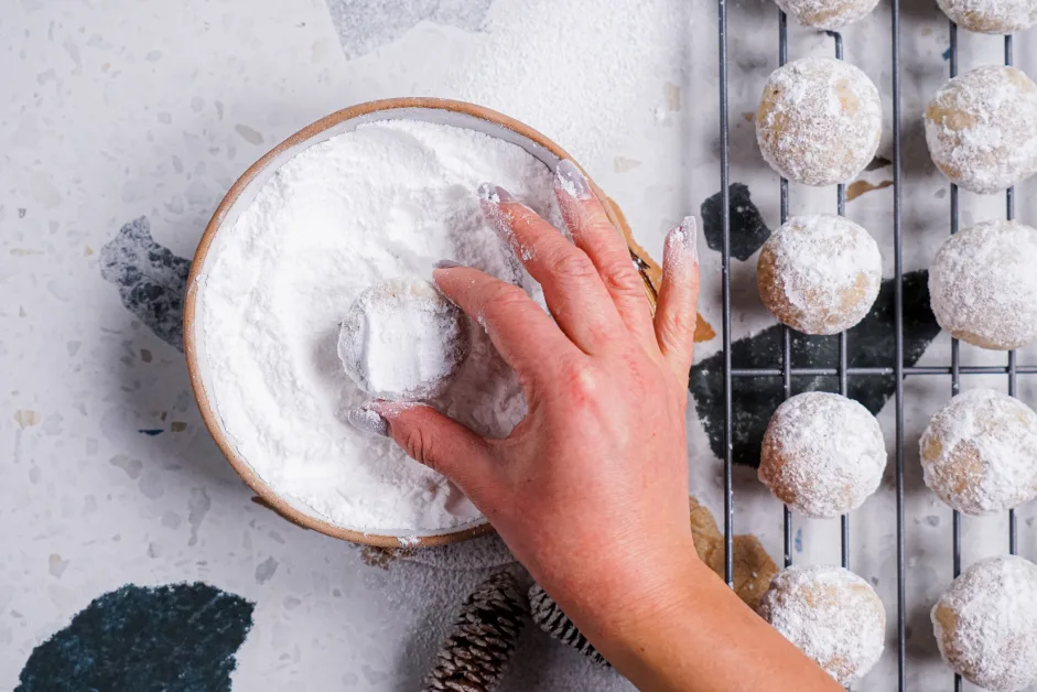 Hands rolling a ball of chocolate cookie dough first in granulated sugar and then in powdered sugar beside two shallow bowls on a kitchen counter