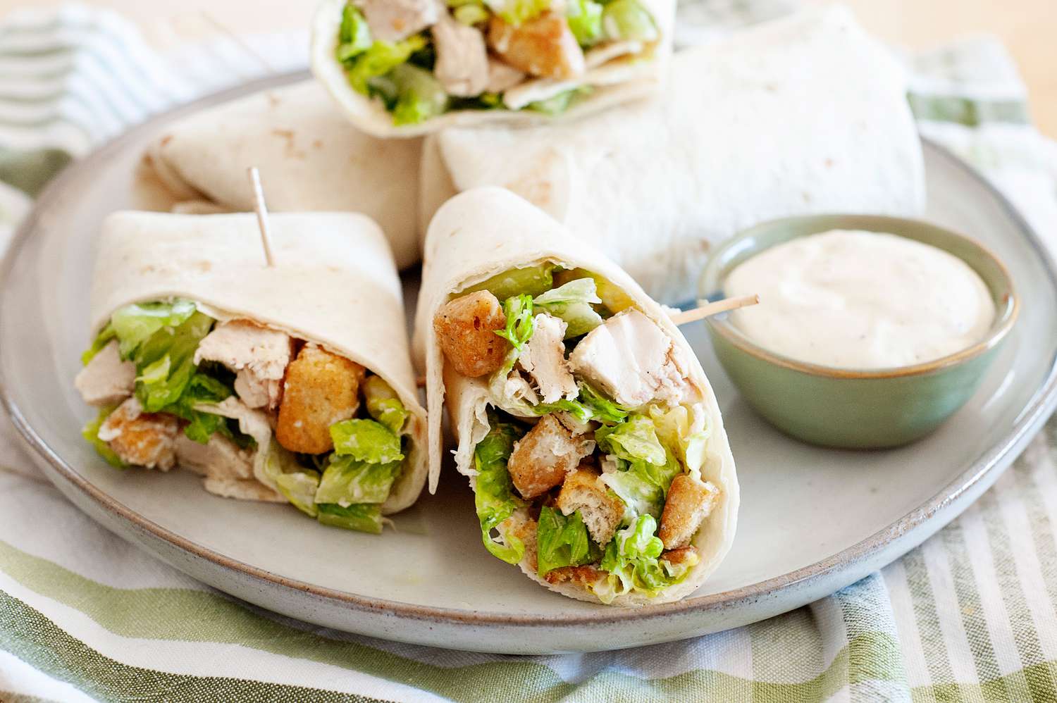 Hands rolling a chicken Caesar wrap tightly on a wooden cutting board with romaine and chicken visible near the edge