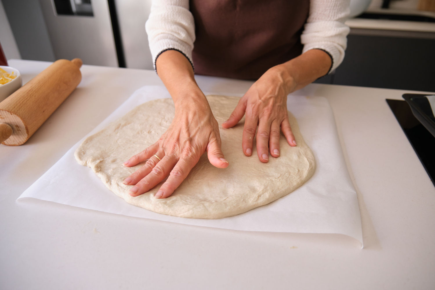 Hands rolling out pizza dough on parchment paper with a wooden rolling pin on a kitchen counter