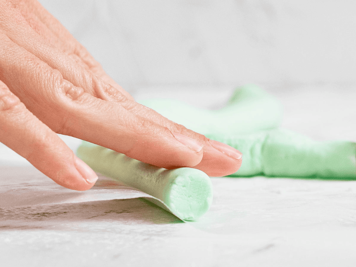 Hands rolling pale green mint dough into a rope on a powdered sugar dusted countertop