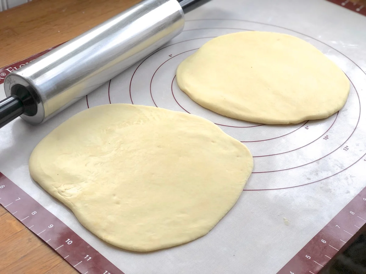 Hands rolling pierogi dough with a wooden rolling pin on a lightly floured countertop, with a partially rolled round and a small dusting of flour nearby