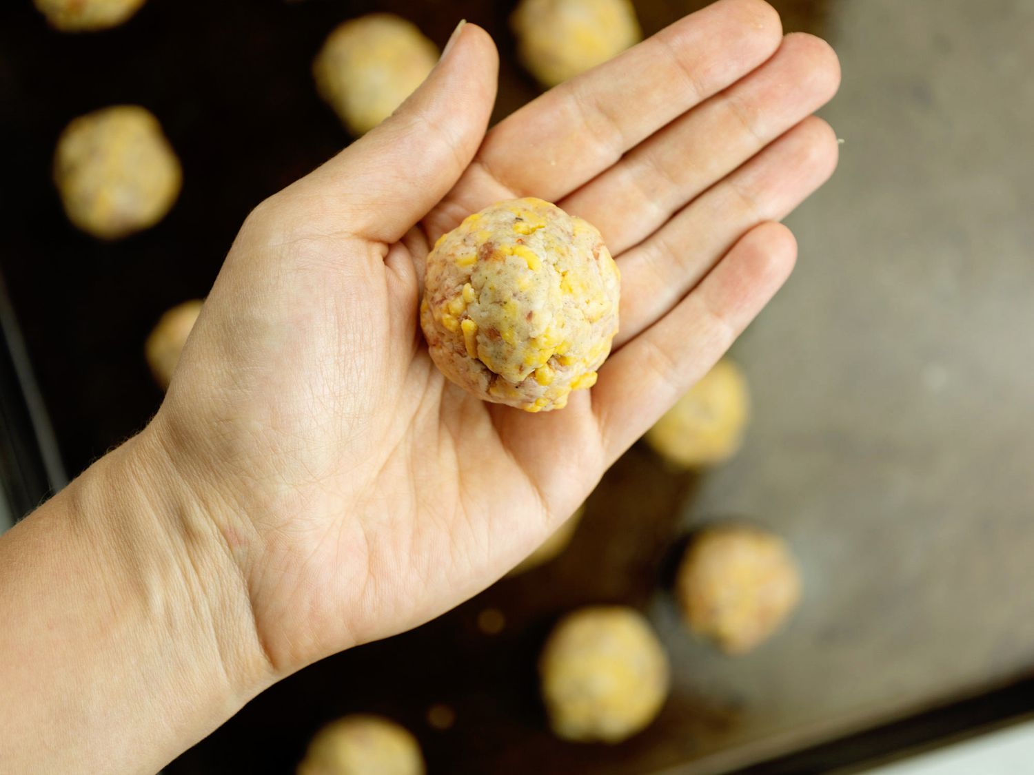 Hands rolling sausage ball mixture into small 1-inch balls over a parchment-lined baking sheet on a kitchen counter