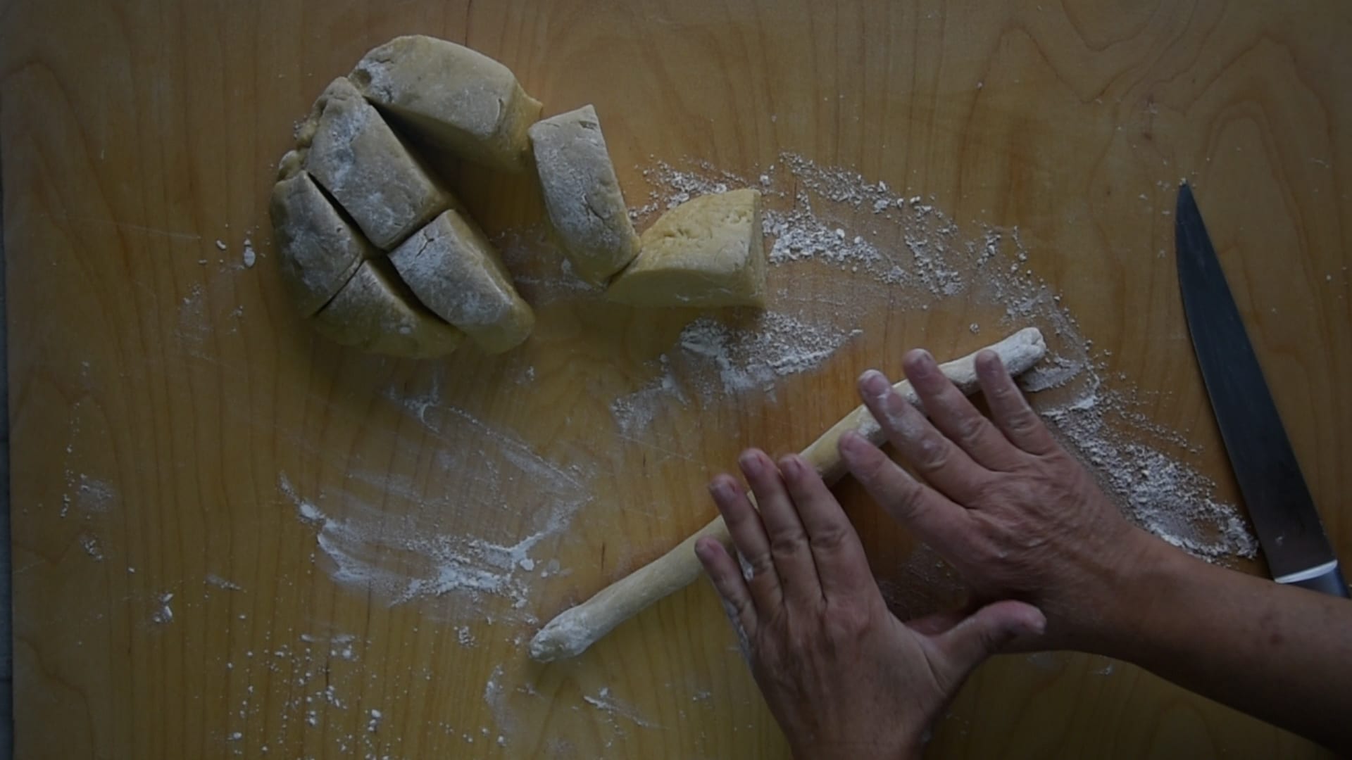 Hands rolling small ropes of struffoli dough on a lightly floured wooden cutting board in a home kitchen