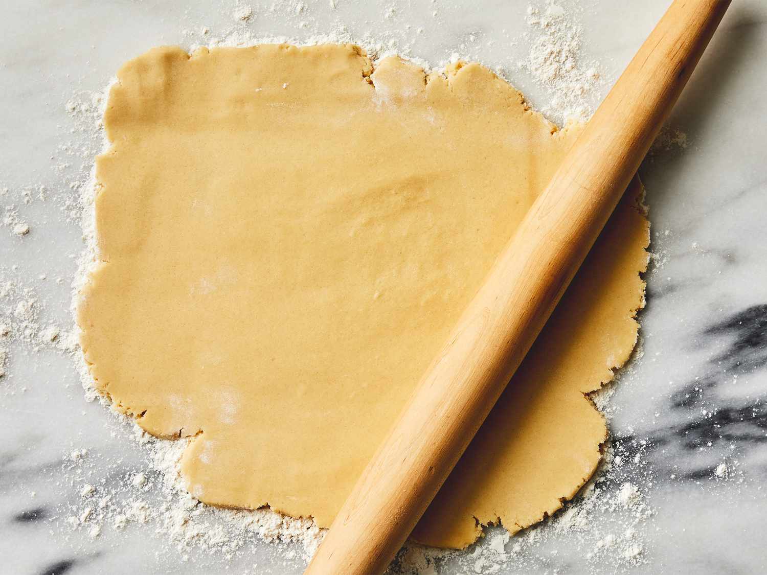 Hands rolling sugar cookie dough on a floured counter with a rolling pin, ready to cut into squares