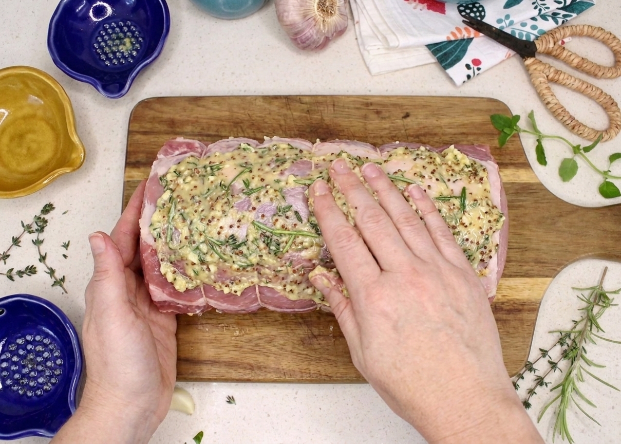 Hands rubbing a garlic herb mustard paste over a raw pork loin roast on a cutting board, kitchen counter in the background