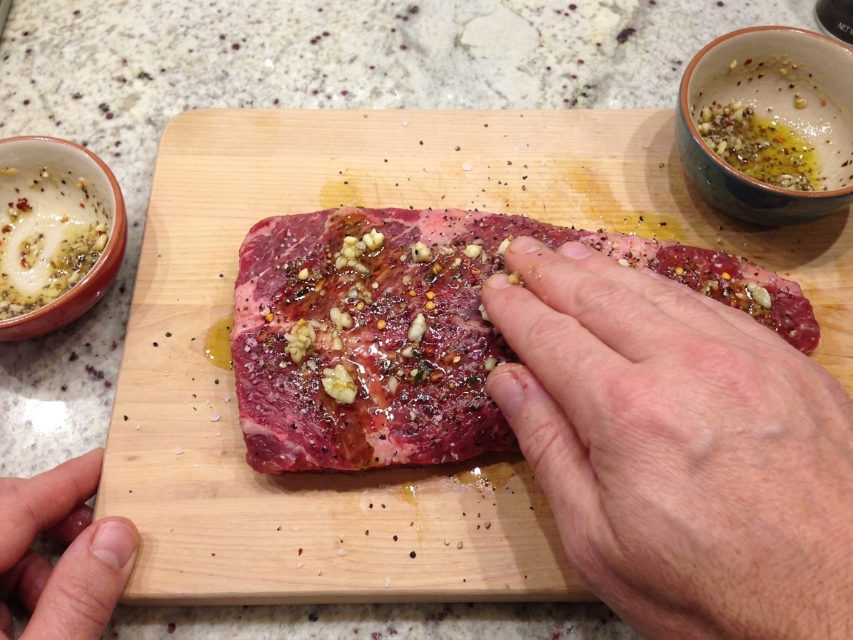 Hands rubbing a seasoned oil and garlic mixture onto a raw flat iron steak on a wooden cutting board in a home kitchen