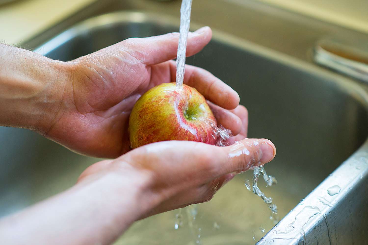 Hands scrubbing apples and carrots under running water at a kitchen sink with a vegetable brush