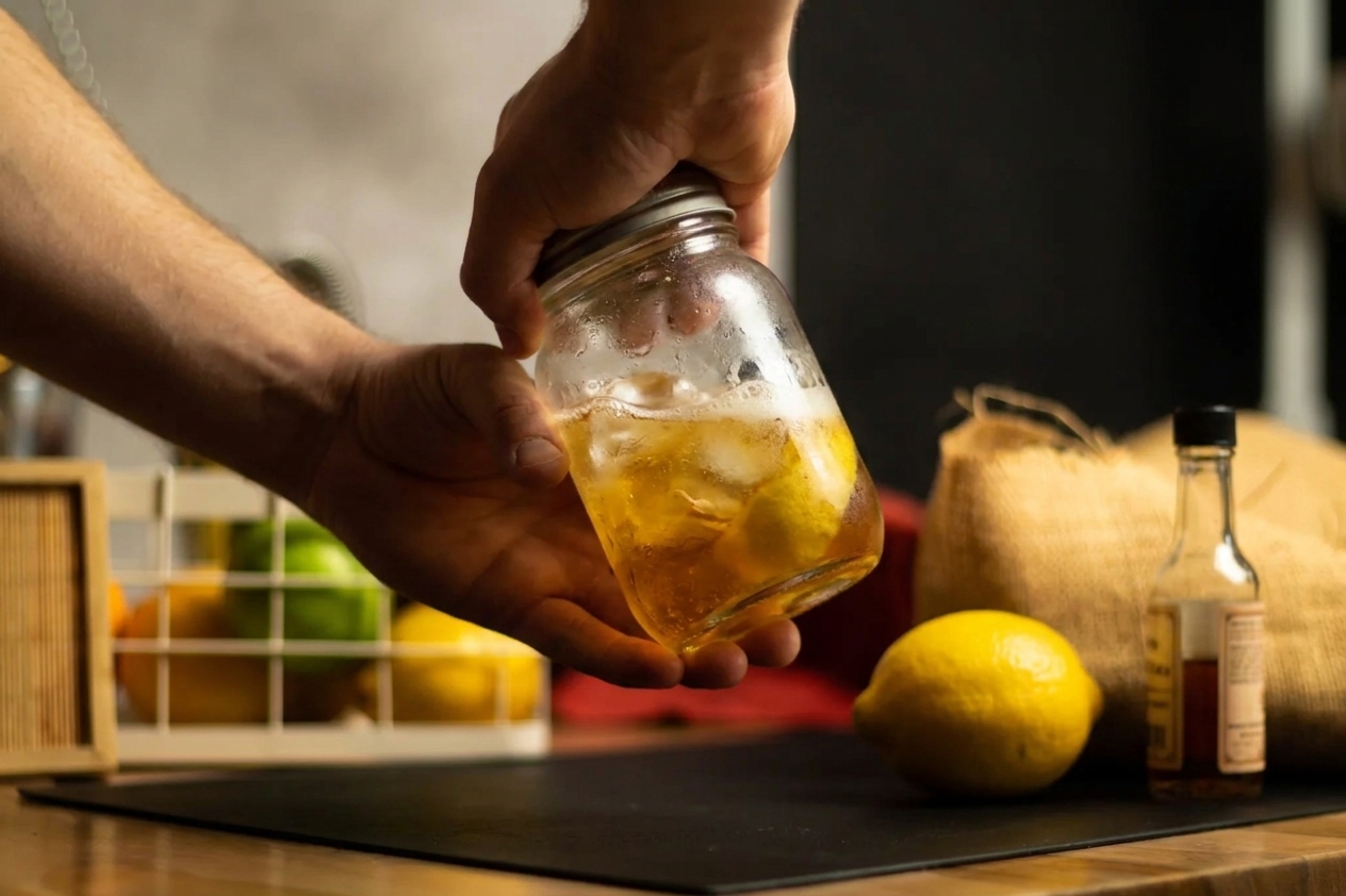 Hands shaking a bourbon cocktail in a mason jar with ice on a kitchen counter, with a lemon and bitters bottle nearby
