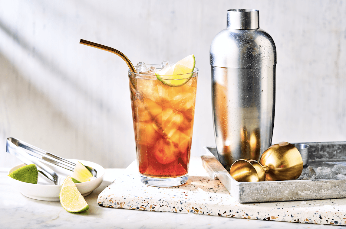 Hands shaking a metal cocktail shaker beside a highball glass packed with ice on a tidy kitchen counter