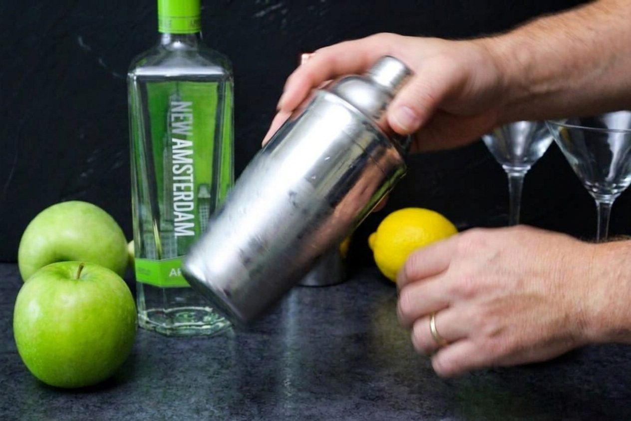 Hands shaking a metal cocktail shaker over a bright kitchen counter with motion blur showing vigorous shaking