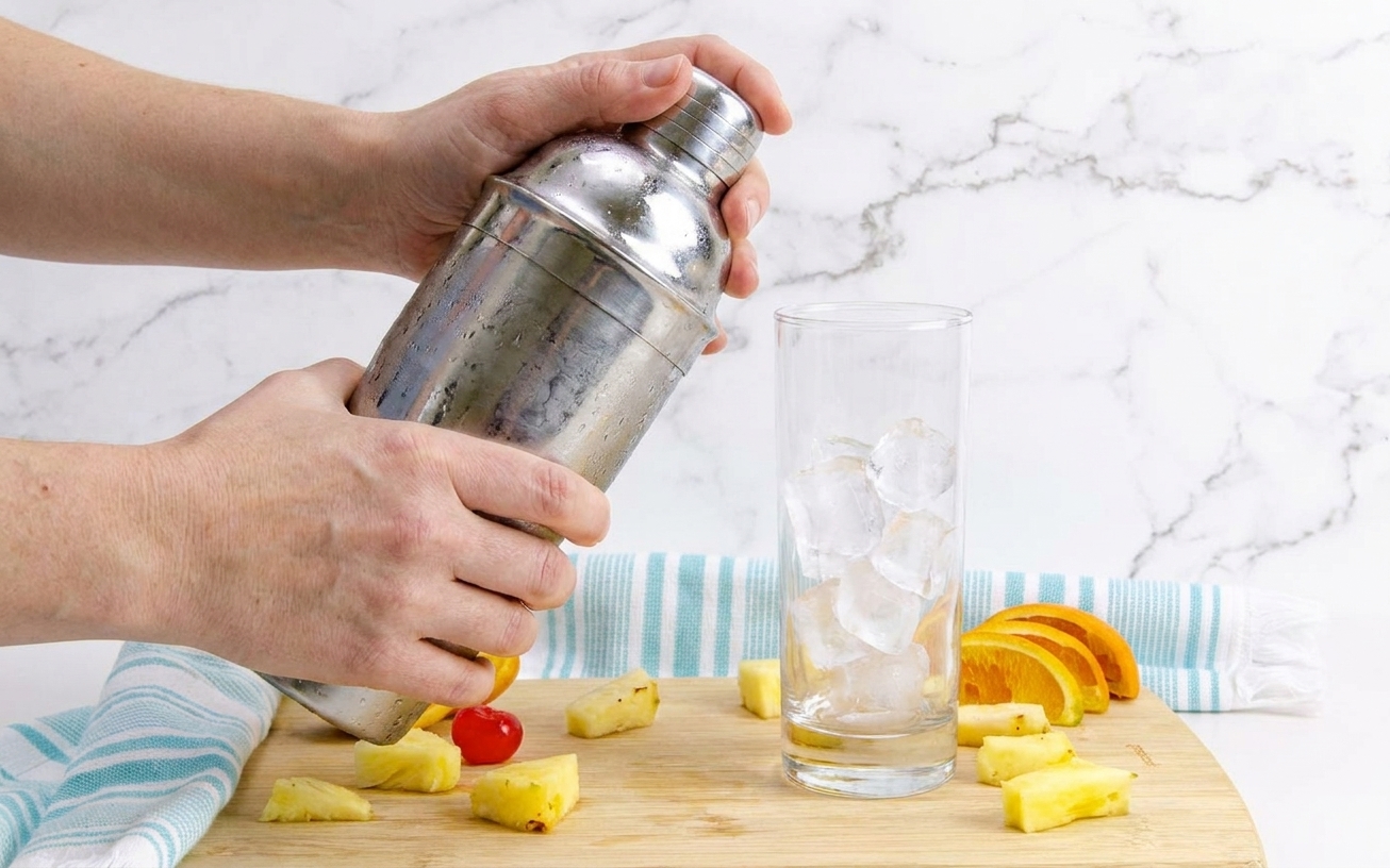 Hands shaking a metal cocktail shaker over a kitchen counter with a highball glass of ice waiting nearby